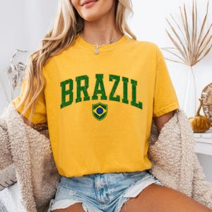 May include: A mustard yellow short-sleeve t-shirt featuring the word BRAZIL in a distressed dark green collegiate font. Below the text is a small shield-shaped emblem displaying the green, yellow, and blue colors of the Brazilian flag.