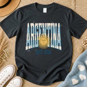 May include: A black short-sleeve t-shirt featuring the word ARGENTINA in large, distressed white and light blue block letters. A golden yellow sun with a human face is centered in the design, with the text EST. 1810 printed in blue below it.