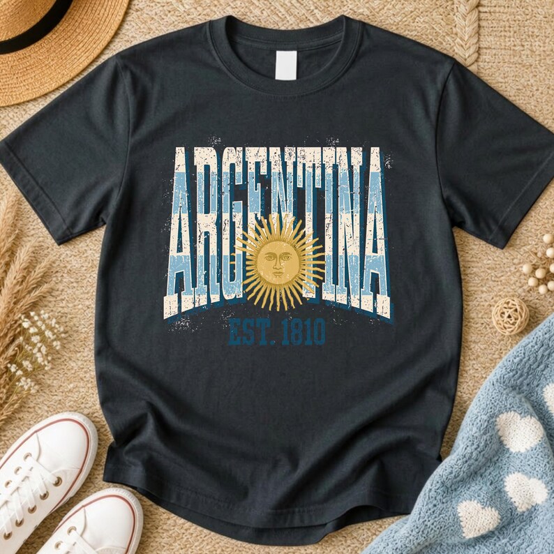 May include: A black short-sleeve t-shirt featuring the word ARGENTINA in large, distressed white and light blue block letters. A golden yellow sun with a human face is centered in the design, with the text EST. 1810 printed in blue below it.