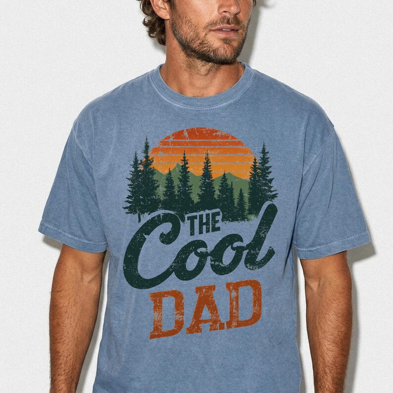 May include: A blue short-sleeve crew neck t-shirt featuring a graphic of a sunset over a forest of pine trees. The text "THE COOL DAD" is printed in dark green and orange lettering below the forest scene. The shirt has a faded, vintage appearance.