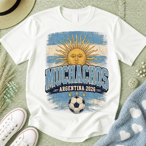 May include: A white short-sleeve t-shirt featuring a distressed graphic of the Argentine sun emblem, a soccer ball, and the text "MUCHACHOS ARGENTINA 2026" in blue and white. The design includes three yellow stars above the soccer ball.