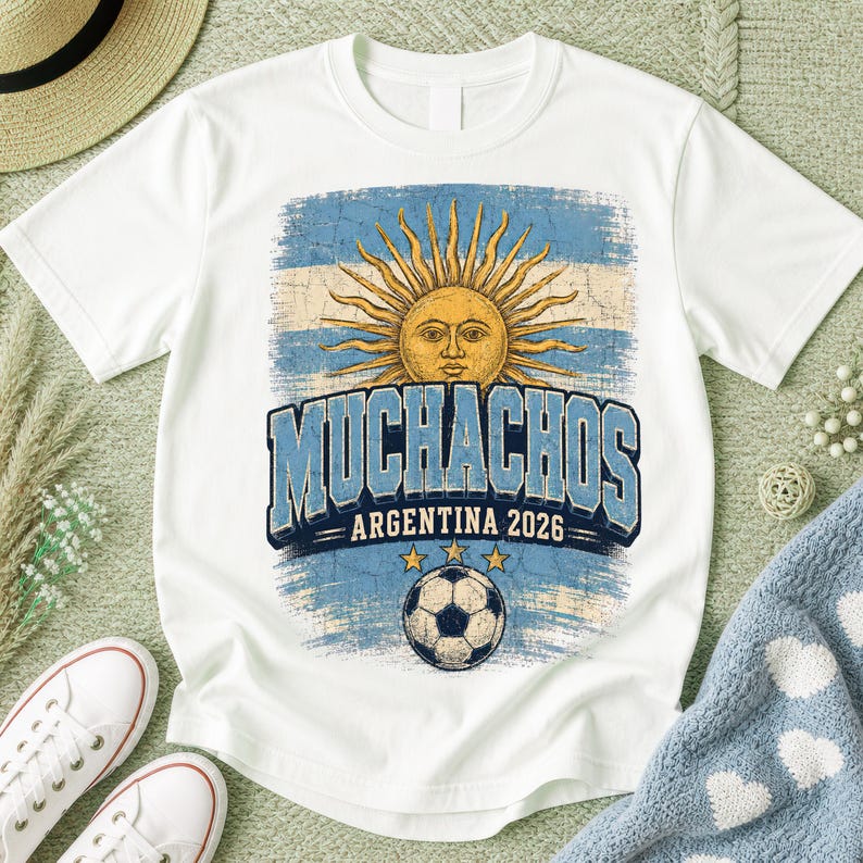 May include: A white short-sleeve t-shirt featuring a distressed graphic of the Argentine sun emblem, a soccer ball, and the text "MUCHACHOS ARGENTINA 2026" in blue and white. The design includes three yellow stars above the soccer ball.
