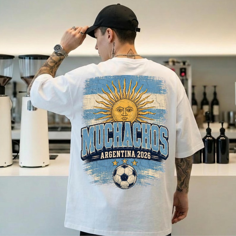 May include: A white short-sleeve t-shirt featuring a distressed graphic on the back. The design includes a yellow sun with a face, the text "MUCHACHOS ARGENTINA 2026" in blue, and a soccer ball graphic over a light blue and white striped background.