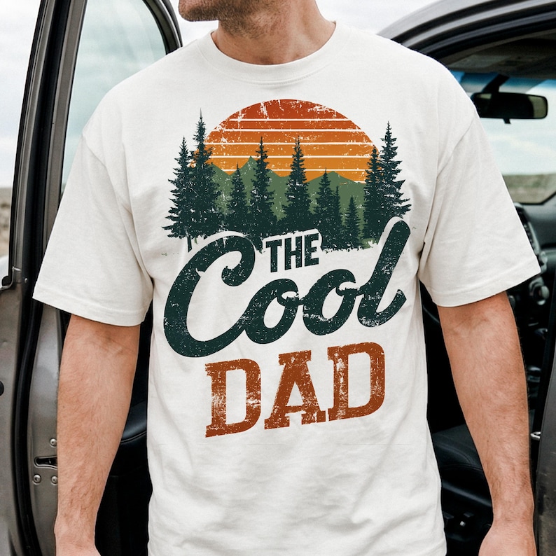May include: Cream-coloured t-shirt with a graphic design featuring a sunset, pine trees, and the words "The Cool Dad" in dark green and orange. The shirt is made of a soft material.