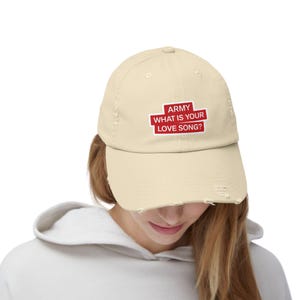 May include: A cream-colored, distressed baseball cap with the text "ARMY WHAT IS YOUR LOVE SONG?" in red and white. The cap has a curved brim and a worn appearance.