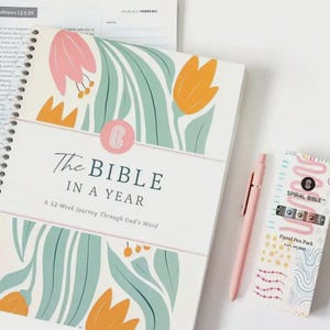 May include: A spiral-bound "The Bible in a Year" journal with floral illustrations in pink, orange, and teal. A pink pen and a "Spiral Bible" pastel pen pack are next to the journal. The journal's cover has the text "The Bible in a Year".
