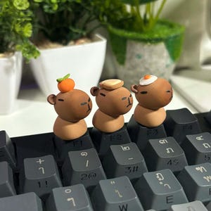 May include: Three small, brown clay figures of capybaras sit on a keyboard. Each capybara has a different food item on its head: an orange, a piece of toast, and a fried egg. The keyboard keys are gray.