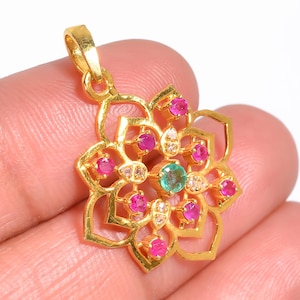 May include: A gold pendant with a floral design. The pendant features a central emerald-coloured stone, surrounded by pink and white gemstones. The pendant is held by a person's fingers.