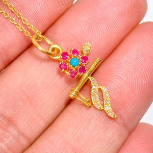 Könnte beinhalten: Goldfarbene Halskette mit Anhänger, die ein Blumendesign mit einem türkisfarbenen Mittelteil und rosa Edelsteinblütenblättern aufweist. Der Anhänger hat auch ein dekoratives Element mit klaren Edelsteinen und hängt an einer zarten Goldkette.