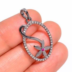 May include: An oval-shaped pendant crafted from dark gunmetal-toned metal, featuring a border encrusted with small clear stones. A stylised flower motif with a single vibrant pink stone at its centre is integrated into the open oval frame.