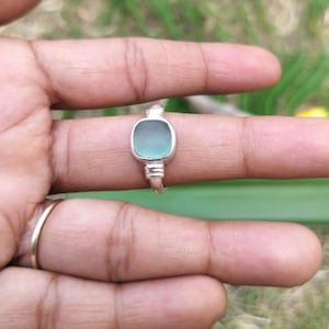 Puede incluir: Un anillo de plata con una piedra de vidrio marino lisa de forma cuadrada en un suave tono azul verdoso. La banda de metal tiene un acabado pulido con un engaste sencillo que asegura la piedra, mostrando un diseño limpio y directo.