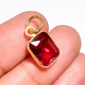 May include: A rectangular pendant featuring a faceted deep red gemstone set in a gold-toned metal bezel. The pendant includes a small circular gold-toned jump ring at the top for attachment, showcasing a polished finish and clear, sharp edges.