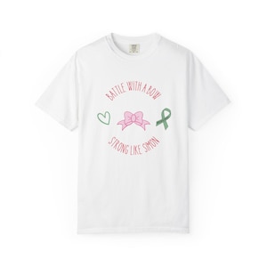 May include: White t-shirt with a crew neck featuring a pink bow, green heart, and green ribbon graphic. The text "BATTLE WITH A BOW STRONG LIKE SIMON" is printed in red around the bow.