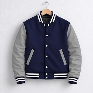 Op de afbeelding: Een marineblauw varsity-jack met lichtgrijze mouwen, een geribbelde kraag met witte strepen, witte drukknopen, twee zijzakken met grijze randen, en geribbelde boorden en zoom met witte strepen op een donkerblauwe ondergrond.