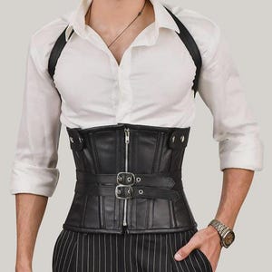 May include: A black corset with a front zipper closure and two horizontal straps featuring silver-toned metal buckles. The corset includes integrated shoulder straps and is worn over a white button-down shirt and black pinstriped trousers.