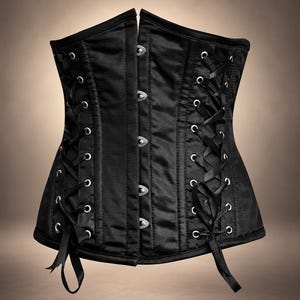 May include: A black corset featuring a front busk closure with silver-toned metal clasps. The garment includes side panels with silver-toned metal eyelets and black ribbon lacing that ties at the bottom, creating a structured, fitted silhouette.