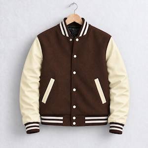 May include: A varsity jacket featuring a dark brown wool-blend body and cream-coloured sleeves. The jacket includes a ribbed collar, cuffs, and hem with white stripes, a front snap-button closure, and two side pockets with cream-coloured trim.