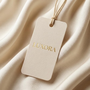 Könnte beinhalten: Ein cremefarbenes rechteckiges Etikett mit dem goldfarbenen Text "LUXORA" wird von einer beigefarbenen Schnur gehalten. Das Etikett befindet sich vor einem Hintergrund aus weichem, fließendem Stoff in einem ähnlichen Cremeton, was eine monochrome Ästhetik erzeugt.