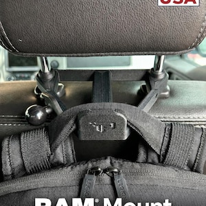 May include: A black plastic car headrest mount featuring a RAM Mount compatible ball joint and a central strap attachment point. The device is secured to the metal headrest posts and supports a black backpack with zippers labelled LEGION.