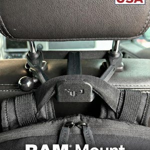 May include: A black plastic car headrest mount featuring a RAM Mount compatible ball joint and a central strap attachment point. The device is secured to the metal headrest posts and supports a black backpack with zippers labeled LEGION.