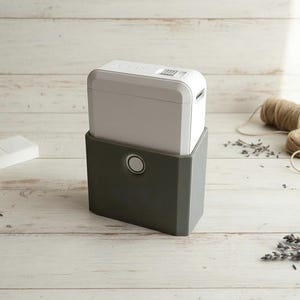 Puede incluir: Una rotuladora blanca y gris se encuentra sobre una base gris oscuro, con un botón circular. Una pequeña suculenta en una maceta blanca, una jabonera blanca y cuerda también están sobre la superficie de madera clara.