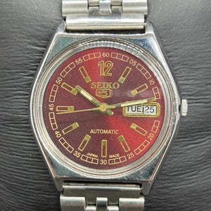 May include: A silver-toned Seiko automatic wristwatch with a burgundy dial. The watch features a day and date display, rectangular hour markers, and a silver-toned metal band. The watch face displays the text "SEIKO" and "AUTOMATIC".