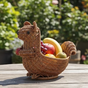 May include: A woven brown squirrel-shaped basket filled with fresh fruit, including a banana, apple, grapes, orange, and kiwi. The basket is made of wicker and sits on a wooden table, with a blurred green background.