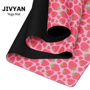 May include: A JIVYAN yoga mat featuring a repeating pattern of red strawberries with green leaves on a light pink background. The mat is partially rolled, revealing a solid black underside with a textured surface finish.