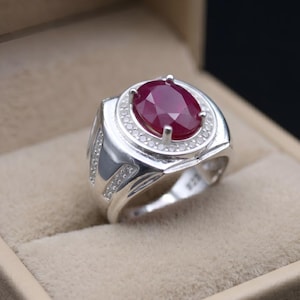 May include: A silver ring featuring a large oval ruby gemstone. The ruby is a deep red hue, encircled by smaller, clear gemstones. The ring is set in a silver band and presented in a beige ring box.