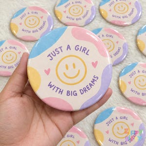 Può includere: Una spilla rotonda con una faccina sorridente gialla, cuori rosa e il testo "JUST A GIRL WITH BIG DREAMS" in viola. Lo sfondo è composto da macchie astratte e ondulate di colore azzurro, rosa tenue, lavanda e giallo pallido.