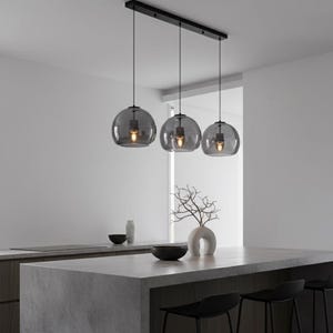 Puede incluir: Tres lámparas colgantes con globos de cristal gris cuelgan de una estructura rectangular negra. Cada globo alberga una bombilla de tono cálido. Las luces están suspendidas sobre una isla de cocina con encimera gris y taburetes negros.