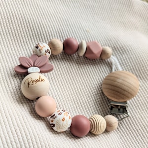 Könnte beinhalten: Ein Schnullerhalter mit runden und geometrischen Perlen in Altrosa, Creme und Naturholz. Das Design umfasst eine Perle mit Blumenmuster, eine große Holzperle mit der Gravur Rosalie sowie einen blumenförmigen Anhänger aus Silikon.
