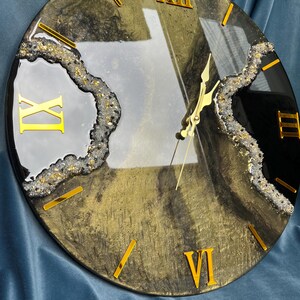 May include: Circular wall clock with a gold and black design. The clock face has gold Roman numerals and hands. The clock has a textured gold and black background with a decorative border.