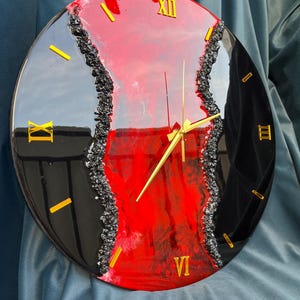Può includere: Orologio da parete rotondo con un design nero e rosso lucido. Il quadrante presenta numeri romani e lancette dorate. Un bordo strutturato grigio scuro separa le sezioni nere e rosse. L'orologio misura circa 30 cm di diametro.