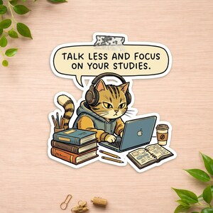 May include: A sticker featuring a brown tabby cat wearing headphones, sitting at a laptop with a stack of books, a coffee cup, and an open notebook. A speech bubble above the cat contains the text: TALK LESS AND FOCUS ON YOUR STUDIES.