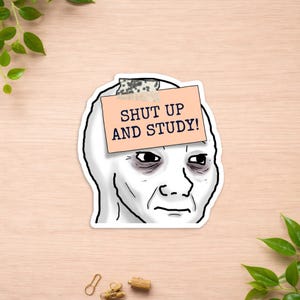 May include: A die-cut sticker featuring a stylized illustration of a tired face with dark circles under the eyes. A light peach-colored rectangular note is taped to the forehead with the text SHUT UP AND STUDY! printed in bold dark blue capital letters.
