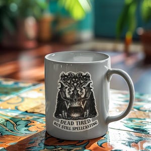 K&ouml;nnte beinhalten: Eine graue Keramiktasse mit einem Aufkleber mit einem Hexenmotiv. Der Aufkleber zeigt drei Hexen um einen Kessel, mit dem Text "DEAD TIRED BUT STILL SPELLCASTING". Die Tasse hat einen gebogenen Henkel.