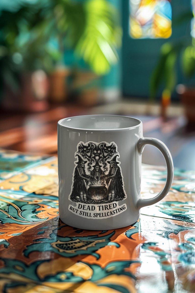 K&ouml;nnte beinhalten: Eine graue Keramiktasse mit einem Aufkleber mit einem Hexenmotiv. Der Aufkleber zeigt drei Hexen um einen Kessel, mit dem Text "DEAD TIRED BUT STILL SPELLCASTING". Die Tasse hat einen gebogenen Henkel.