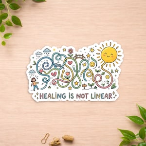 May include: A colorful sticker with the text "HEALING IS NOT LINEAR." The design features a maze-like path with a person, a broken heart, flowers, stars, and a smiling sun. The sticker is on a light wood surface.