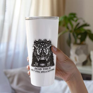 K&ouml;nnte beinhalten: Ein wei&szlig;er Becher mit einer schwarz-wei&szlig;en Grafik von drei Skelettfiguren um einen Kessel. Der Text "DEAD TIRED BUT STILL SPELLCASTING" befindet sich unter der Grafik. Der Becher wird von einer Person gehalten.