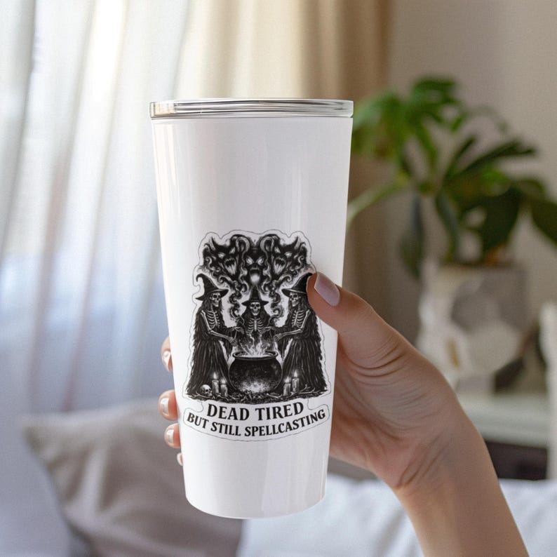 K&ouml;nnte beinhalten: Ein wei&szlig;er Becher mit einer schwarz-wei&szlig;en Grafik von drei Skelettfiguren um einen Kessel. Der Text "DEAD TIRED BUT STILL SPELLCASTING" befindet sich unter der Grafik. Der Becher wird von einer Person gehalten.