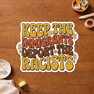 May include: A die-cut sticker featuring the text KEEP THE IMMIGRANTS DEPORT THE RACISTS in a retro, wavy font. The lettering uses a color palette of mustard yellow, burnt orange, and dark brown, accented with small sunburst and peace sign icons.
