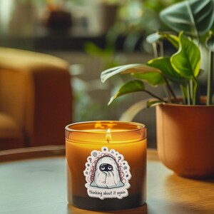 May include: A lit candle in a glass jar with a sticker that says "thinking about it again." The candle has a cartoon ghost design. The candle sits on a wooden table next to a potted plant with green leaves.