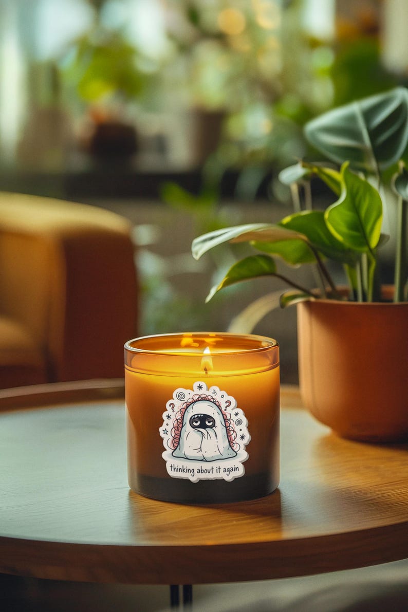 May include: A lit candle in a glass jar with a sticker that says "thinking about it again." The candle has a cartoon ghost design. The candle sits on a wooden table next to a potted plant with green leaves.