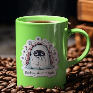 May include: A bright green ceramic coffee mug filled with dark coffee, with steam rising from the top. The mug features a sticker with a cartoon ghost and the text "thinking about it again". The mug is surrounded by coffee beans.