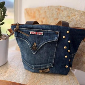 May include: A denim tote bag with a front pocket and brown leather handles. The bag features a zipper closure, brass studs, and a small rectangular patch with red and white text. The bag is sitting on a stone surface.