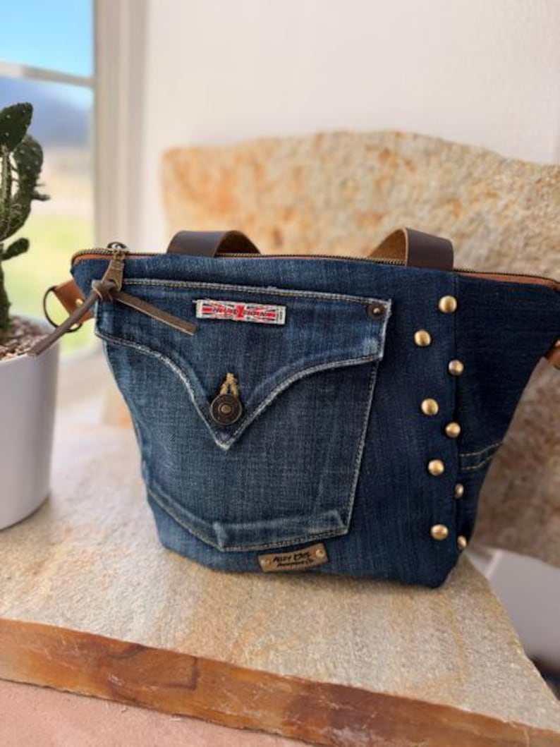 May include: A denim tote bag with a front pocket and brown leather handles. The bag features a zipper closure, brass studs, and a small rectangular patch with red and white text. The bag is sitting on a stone surface.