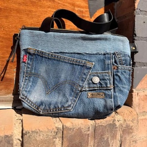 May include: A denim handbag with a black leather strap. The bag features a front pocket from a pair of Levi's jeans, complete with the iconic red tag. The bag has a zipper closure and a small metal tag.