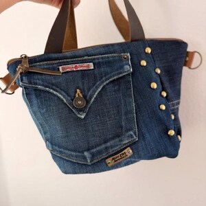 May include: A denim tote bag with brown leather handles and accents. The bag features a front pocket with a button closure and gold-colored studs along the side. The bag has a zipper closure and a small metal tag that reads "Alley Cats Apparel Co."