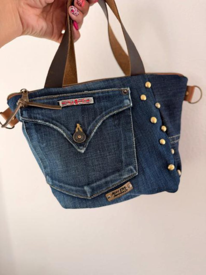 May include: A denim tote bag with brown leather handles and accents. The bag features a front pocket with a button closure and gold-colored studs along the side. The bag has a zipper closure and a small metal tag that reads "Alley Cats Apparel Co."